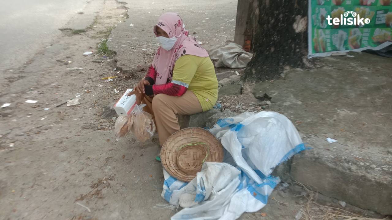 Haimah yang mengenakan kerudung merah muda sambil memegang tissu dan kerupuk opak untuk dijajakan, sedang duduk di bawah pohon menunggu lampu merah menyala. Foto: Nadwa Rifada/Telisik
